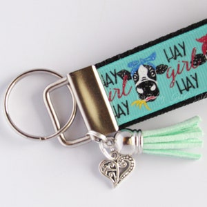 May include: A turquoise and black keychain with a cow design and the text "Hay Girl Hay". The keychain has a silver key ring and a light green tassel.