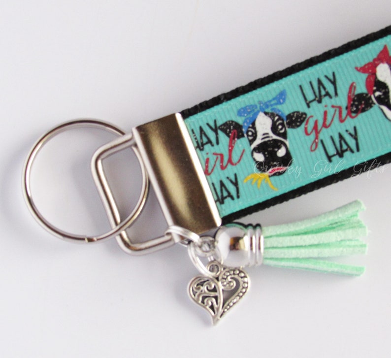 May include: A turquoise and black keychain with a cow design and the text "Hay Girl Hay". The keychain has a silver key ring and a light green tassel.
