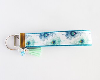 May include: A teal blue and white key chain with a peacock feather design. The key chain has a silver key ring and a light green tassel.