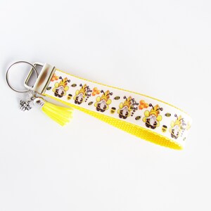 May include: Yellow and white key chain with a bee gnome design. The key chain has a silver key ring and a yellow tassel. The ribbon has the text "Bee Kind" printed on it.