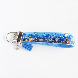 May include: A blue and white key chain with a seashell charm and a blue tassel. The key chain features a colorful underwater scene with fish and coral.