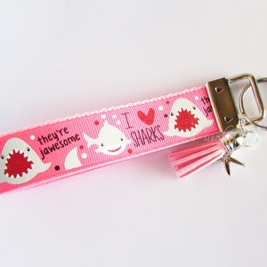 K&ouml;nnte beinhalten: Rosafarbener Schl&uuml;sselanh&auml;nger mit einem wei&szlig;en Hai-Design und dem Text "Ich liebe Haie, sie sind genial". Der Schl&uuml;sselanh&auml;nger hat einen rosa Quastenanh&auml;nger und einen silbernen Seestern-Anh&auml;nger.
