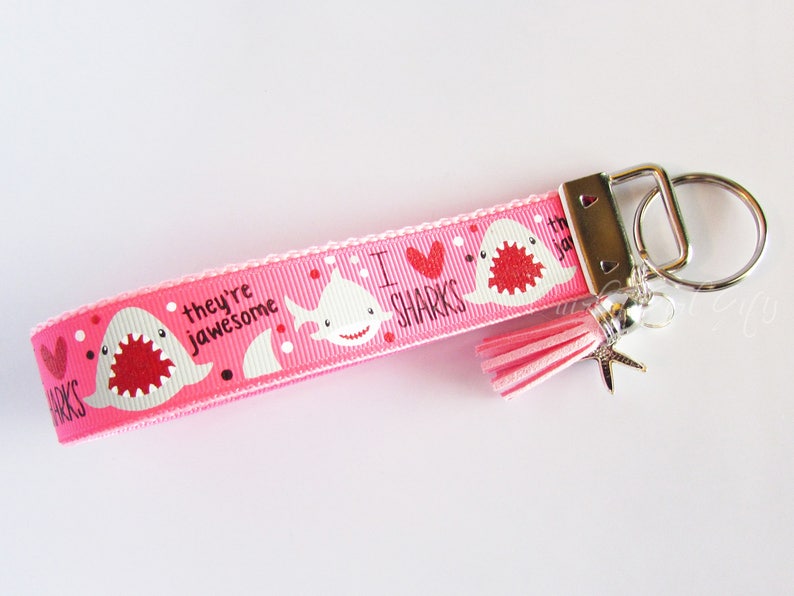 K&ouml;nnte beinhalten: Rosafarbener Schl&uuml;sselanh&auml;nger mit einem wei&szlig;en Hai-Design und dem Text "Ich liebe Haie, sie sind genial". Der Schl&uuml;sselanh&auml;nger hat einen rosa Quastenanh&auml;nger und einen silbernen Seestern-Anh&auml;nger.