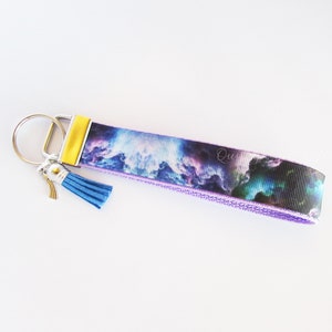 May include: A purple and blue key chain with a galaxy print and a blue tassel. The key chain has a silver key ring and a yellow accent.