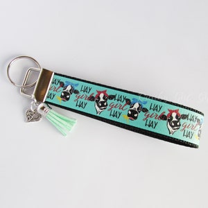 May include: A black and turquoise key chain with a cow print design and the text "Hay Girl Hay" repeated. The key chain has a silver heart charm and a turquoise tassel.
