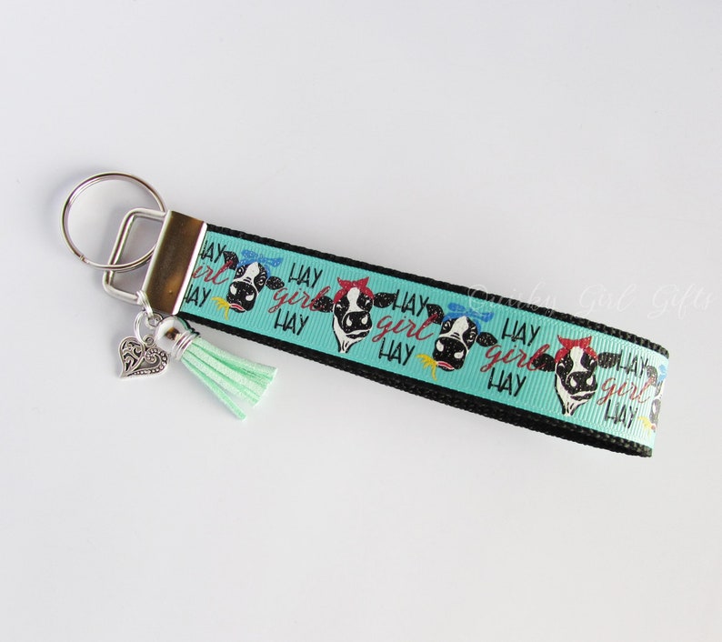 May include: A black and turquoise key chain with a cow print design and the text "Hay Girl Hay" repeated. The key chain has a silver heart charm and a turquoise tassel.