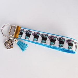 May include: A keychain with a blue and white patterned strap featuring coffee cup graphics. The keychain has a silver-coloured ring and a charm of a teacup. A blue tassel hangs from the strap.