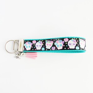 May include: A turquoise blue key chain with a black ribbon featuring a repeating pattern of white sugar skulls with pink and purple flowers. The ribbon has white polka dots. A pink tassel and a silver skull charm are attached to the key chain.