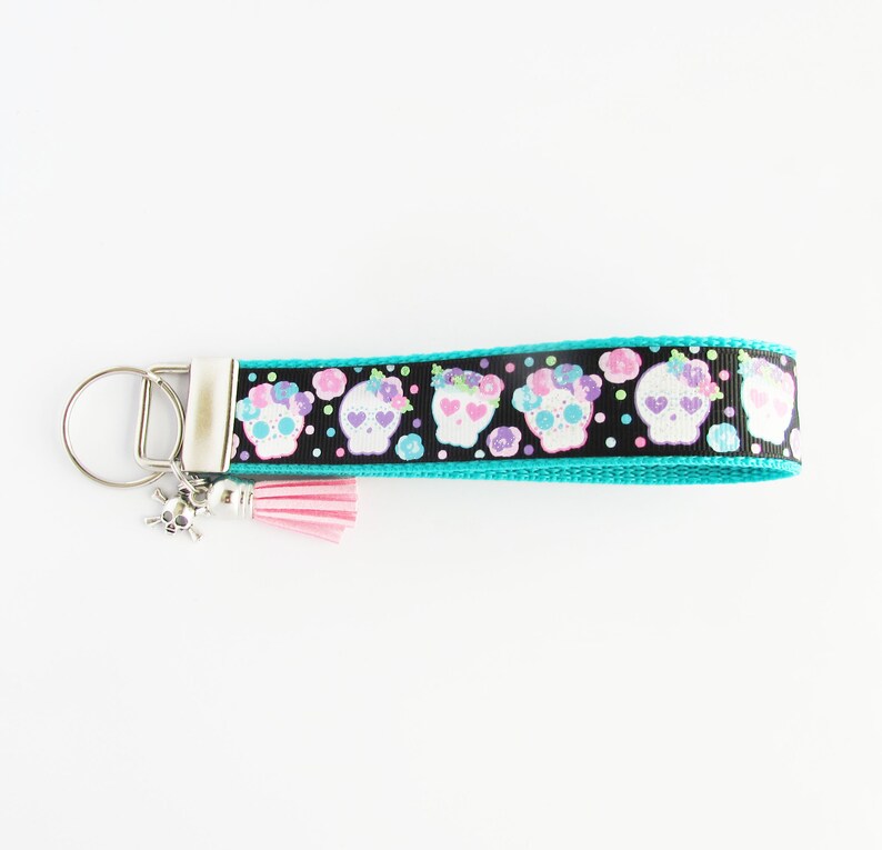 May include: A turquoise blue key chain with a black ribbon featuring a repeating pattern of white sugar skulls with pink and purple flowers. The ribbon has white polka dots. A pink tassel and a silver skull charm are attached to the key chain.