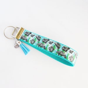 May include: A turquoise blue key chain with a silver seashell charm and a blue tassel. The key chain features a repeating pattern of gray sea turtles on a white background.