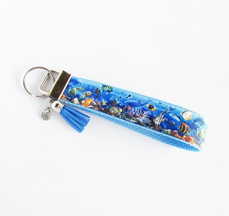 May include: A blue key chain with a seashell charm and a tassel. The key chain features a colorful print of tropical fish and coral.