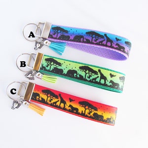 May include: Three colorful keychains with a black silhouette design of African animals and trees. The keychains are labeled A, B, and C. The keychains are purple, green, and orange.