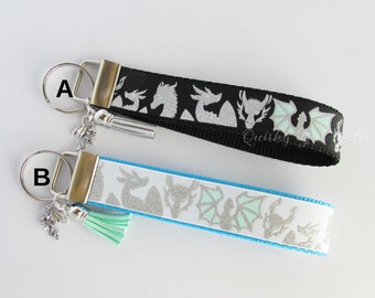 May include: Two keychains with silver dragon silhouettes on black and blue ribbon. The keychain on the left has a silver charm and a black tassel. The keychain on the right has a silver charm and a light blue tassel.  The text "Quirky" is visible on the black ribbon.