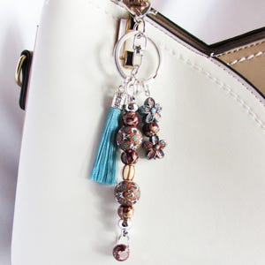 May include: A bag charm featuring a silver-tone metal keyring and lobster clasp. The charm includes a light blue fabric tassel, brown patterned beads, a striped bead, and two small dark teal flower-shaped beads hanging from silver jump rings.