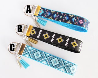 May include: Three keychains with different geometric patterns. Each keychain has a gold-colored key ring, a blue tassel, and a silver feather charm. The keychains are made of black, blue, and white fabric.