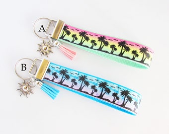 May include: Two keychains with a palm tree and sunset design. Keychain A is green with a pink tassel. Keychain B is blue with a turquoise tassel. Both keychains have a silver sun charm.
