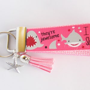 K&ouml;nnte beinhalten: Rosafarbener Schl&uuml;sselanh&auml;nger mit einem silbernen Schl&uuml;sselring, einem rosa Quastenanh&auml;nger und einem silbernen Seestern-Anh&auml;nger. Der Schl&uuml;sselanh&auml;nger hat ein rosa Band mit einer wei&szlig;en Hai-Grafik und dem Text "they're awesome" und "I  SHARK".