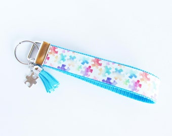 May include: A blue and white keychain with a rainbow puzzle piece pattern and a blue tassel. The keychain has a silver key ring and a silver puzzle piece charm.