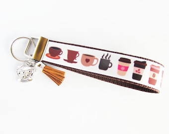 May include: A white wristlet keychain with a brown border and coffee cup designs. The keychain has a silver charm of a coffee pot and a brown tassel. The coffee cup designs include various colors and styles.