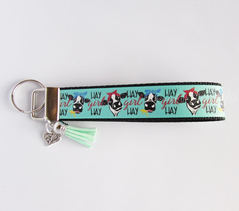 May include: A turquoise blue and black key chain with a cow print design and the text "Hay Girl Hay". The key chain has a silver charm in the shape of a heart and a turquoise tassel.