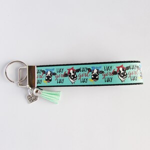 May include: A turquoise and black keychain with a cow print design and the text "Hay Girl Hay" repeated. The keychain has a silver key ring and a silver charm in the shape of a heart. A turquoise tassel hangs from the bottom of the keychain.