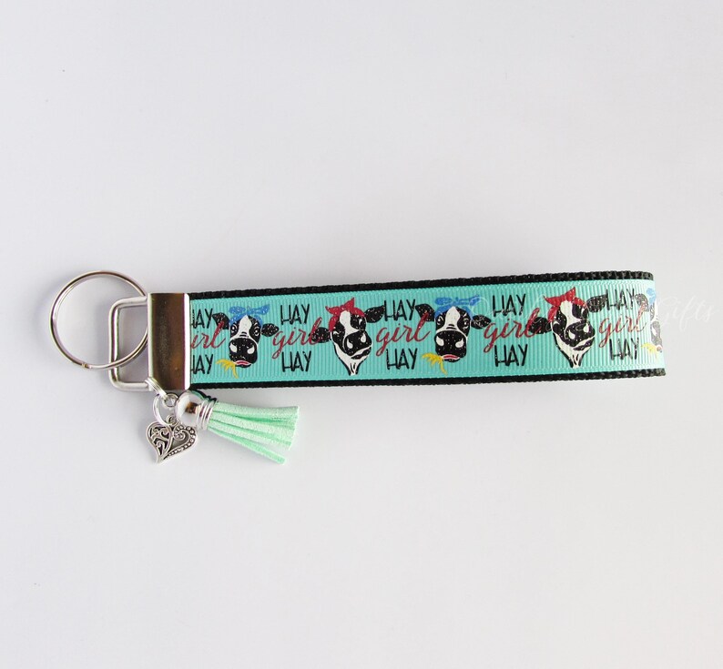 May include: A turquoise and black keychain with a cow print design and the text "Hay Girl Hay" repeated. The keychain has a silver key ring and a silver charm in the shape of a heart. A turquoise tassel hangs from the bottom of the keychain.