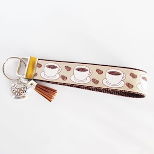 May include: A coffee-themed keyring with a beige strap patterned with coffee cups and beans. It has a silver coffee cup charm and a brown tassel. The keyring is a perfect accessory for coffee enthusiasts.