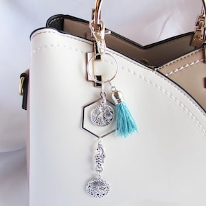 May include: A silver-toned keychain with a teal tassel and ocean-themed charms, including a seahorse and a sand dollar. The keychain is attached to a white and beige handbag with gold hardware.