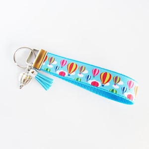 May include: A blue key chain with a silver hot air balloon charm and a blue tassel. The key chain is decorated with a pattern of colorful hot air balloons against a light blue sky.