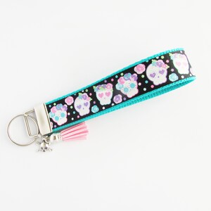 May include: A turquoise and black key chain with a pink tassel. The key chain features a repeating pattern of colorful sugar skulls with flowers. A silver skull charm is attached to the key ring.