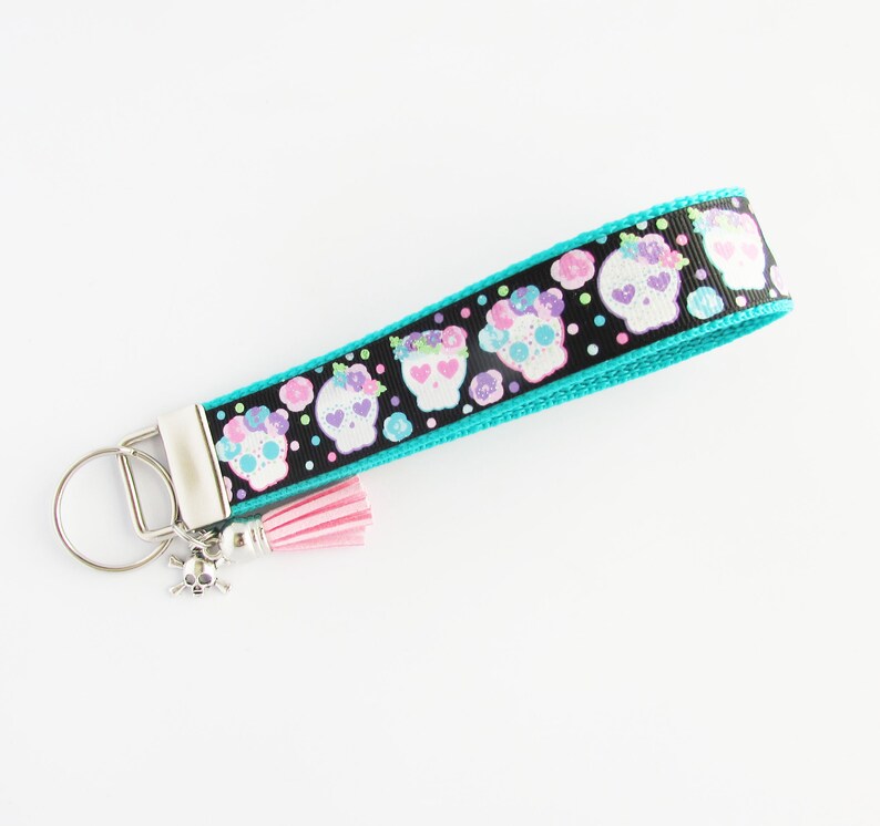 May include: A turquoise and black key chain with a pink tassel. The key chain features a repeating pattern of colorful sugar skulls with flowers. A silver skull charm is attached to the key ring.