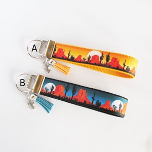 May include: Two keychains with desert landscape designs. One keychain has a yellow and orange sunset background, the other has a blue and black night sky. Each keychain has a metal ring, a tassel, and a cactus charm.