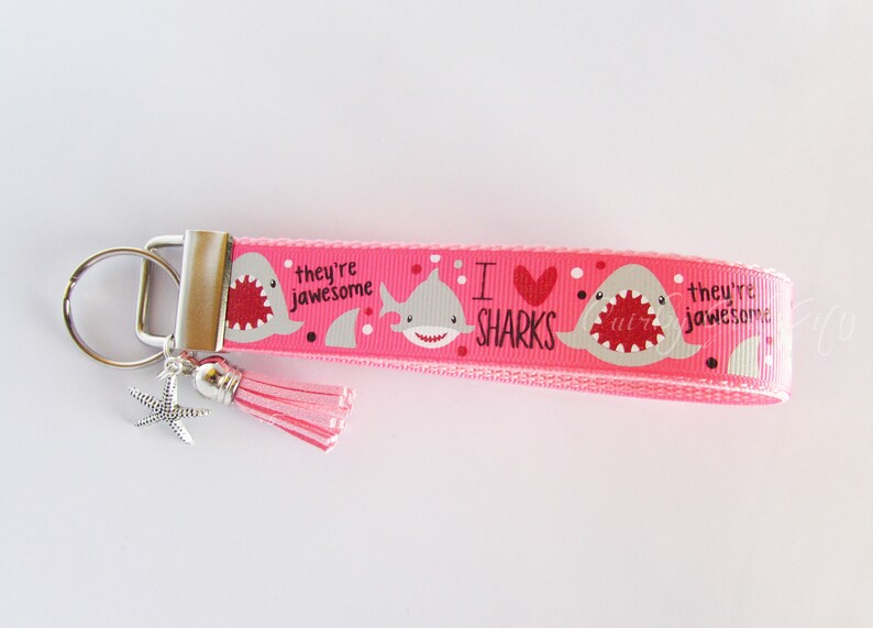 K&ouml;nnte beinhalten: Rosafarbener Schl&uuml;sselanh&auml;nger mit einem Hai-Design und dem Text "Ich liebe Haie, sie sind gro&szlig;artig". Der Schl&uuml;sselanh&auml;nger hat einen silbernen Schl&uuml;sselring und einen rosa Quasten.