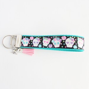 May include: A turquoise and black key chain with a pink tassel and a silver skull charm. The key chain features a repeating pattern of white sugar skulls with pink and purple flowers.