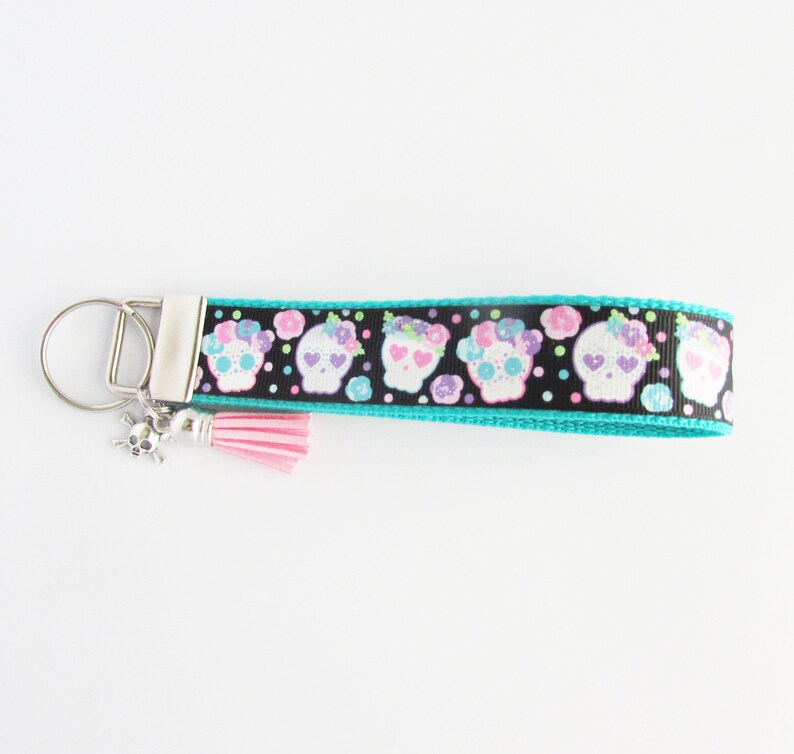 May include: A turquoise and black key chain with a pink tassel and a silver skull charm. The key chain features a repeating pattern of white sugar skulls with pink and purple flowers.