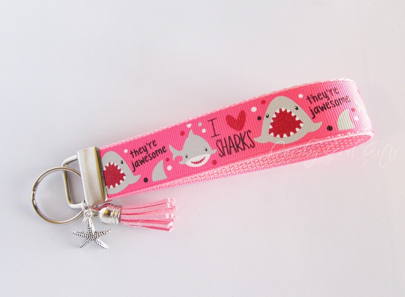 K&ouml;nnte beinhalten: Rosafarbener Schl&uuml;sselanh&auml;nger mit einem Hai-Design und dem Text "Ich liebe Haie, sie sind genial". Der Schl&uuml;sselanh&auml;nger hat einen silbernen Schl&uuml;sselring und einen rosa Quasten.