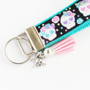May include: A turquoise and black key chain with a pink tassel and a silver skull charm. The key chain features a repeating pattern of colorful sugar skulls.
