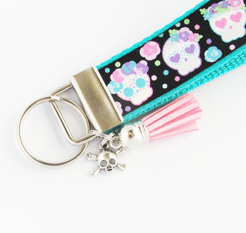 May include: A turquoise and black key chain with a pink tassel and a silver skull charm. The key chain features a repeating pattern of colorful sugar skulls.