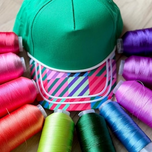 May include: A bright green baseball cap with a white trim and a multi-colored plaid brim featuring pink, purple, blue, and green diagonal stripes, surrounded by an array of eleven colorful embroidery thread spools in various vibrant shades.