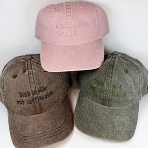 May include: Three baseball caps in pink, brown, and olive green. The pink cap features the embroidered text "coffee walk". The brown and olive green caps both feature the embroidered text "dead inside but caffeinated" in a dark font.