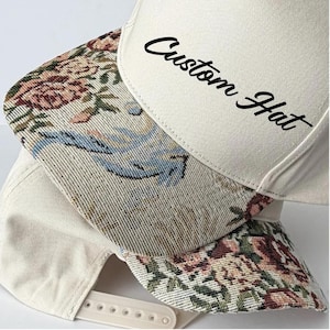May include: A cream-colored baseball cap with a snapback closure and a woven floral tapestry brim featuring pink roses and blue accents. The front panel of the cap displays the black text Custom Hat in a cursive script font, centered on the fabric.