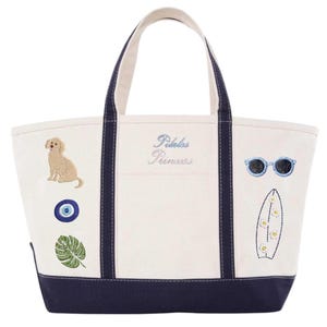 May include: Cream-colored tote bag with navy blue accents and handles. Embroidered with a golden retriever, sunglasses, a surfboard, and the words "Pilates Princess" in light blue. Features a navy blue bottom.