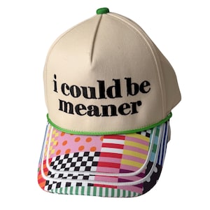 May include: A beige baseball cap featuring the black embroidered text i could be meaner on the front panel. The cap includes a bright green cord trim above the brim, which is decorated with a colorful patchwork pattern of dots, stripes, and checks.