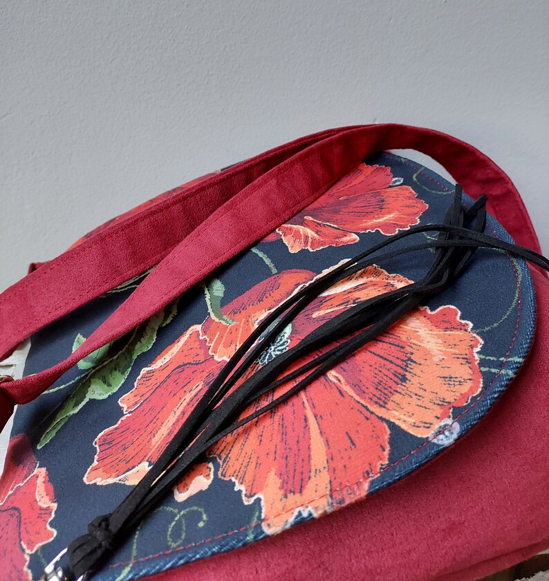 May include: A burgundy crossbody bag with a flap closure. The flap is made of dark blue fabric with a floral print of red poppies. The bag has a black tassel.