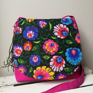 May include: A vibrant, floral-patterned shoulder bag. The bag features a black fabric adorned with colorful flowers in shades of pink, blue, yellow, and purple. The base and strap are a solid magenta color. The bag has a flap closure and a long strap.