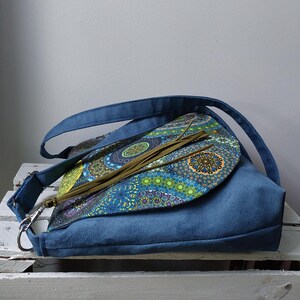May include: A blue suede shoulder bag with a flap featuring a colorful, patterned design. The bag has a long strap and a silver-colored clasp. The flap is adorned with long, olive-green tassels. The bag is sitting on a white surface.