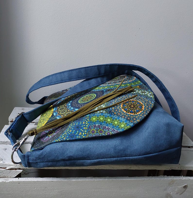 May include: A blue suede shoulder bag with a flap featuring a colorful, patterned design. The bag has a long strap and a silver-colored clasp. The flap is adorned with long, olive-green tassels. The bag is sitting on a white surface.