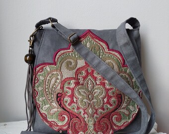 Pode incluir: Uma bolsa de ombro de lona cinza com um grande desenho floral bordado em tons de vermelho, verde e dourado. A bolsa tem uma alça longa ajustável.