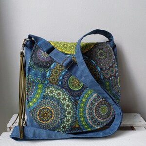 May include: A blue crossbody bag with a colorful mandala pattern. The bag features a blue adjustable strap, a flap closure, and a decorative olive green tassel. The mandala design incorporates shades of blue, green, yellow, and purple.