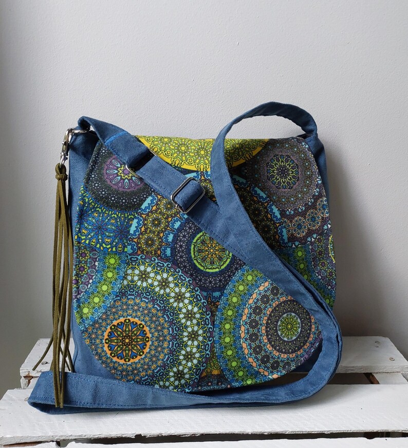 May include: A blue crossbody bag with a colorful mandala pattern. The bag features a blue adjustable strap, a flap closure, and a decorative olive green tassel. The mandala design incorporates shades of blue, green, yellow, and purple.